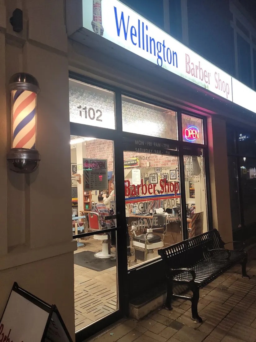 Wellington Barbershop storefront at night with the lit blue sign and a barber pole